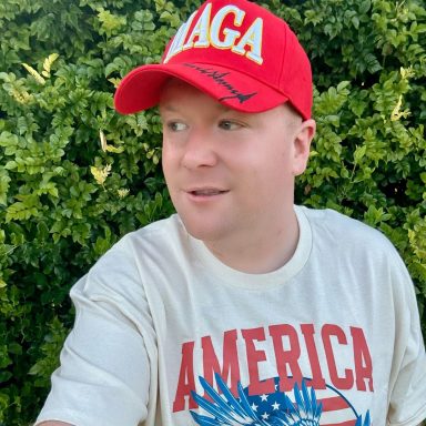Man wearing a red cap and a white "America" t-shirt in front of greenery.